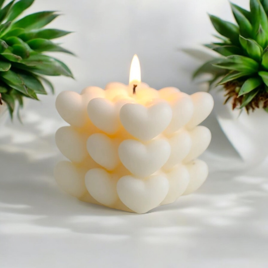 Lit white heart cube scented candle with a warm glow beside green plants