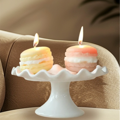Two lit macaron scented candles in yellow and peach on a white cake stand